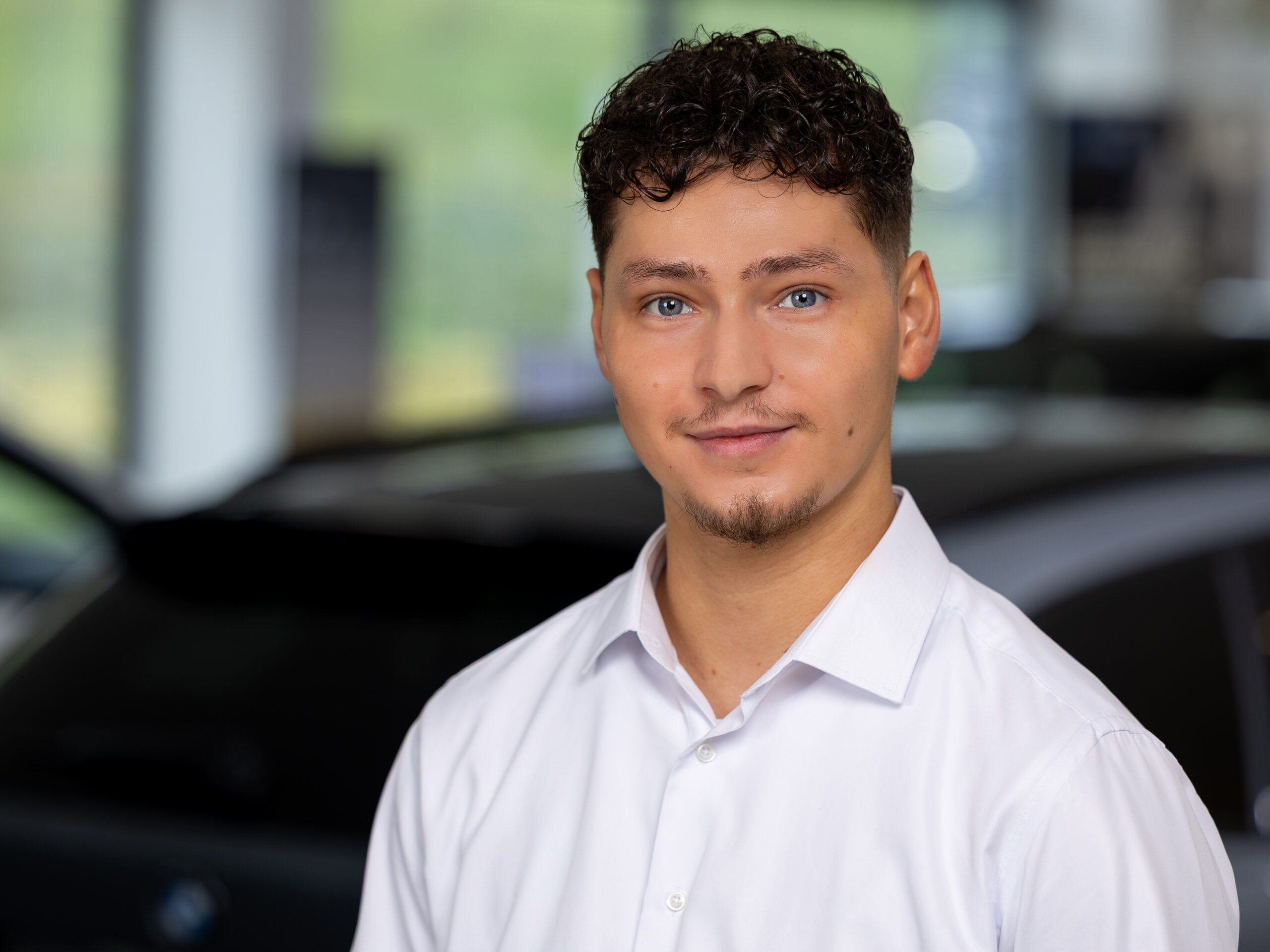Kaltenbach Gruppe – Ein junger Mann mit kurzen lockigen Haaren und blauen Augen, der ein weißes Hemd trägt, lächelt in die Kamera. Der unscharfe Hintergrund deutet auf ein Auto und eine helle Innenumgebung hin, was auf eine moderne Atmosphäre in Kaltenbach Remscheid hindeutet. – BMW, MINI, KIA, BMW Motorrad & Honda Motorrad Partner in NRW
