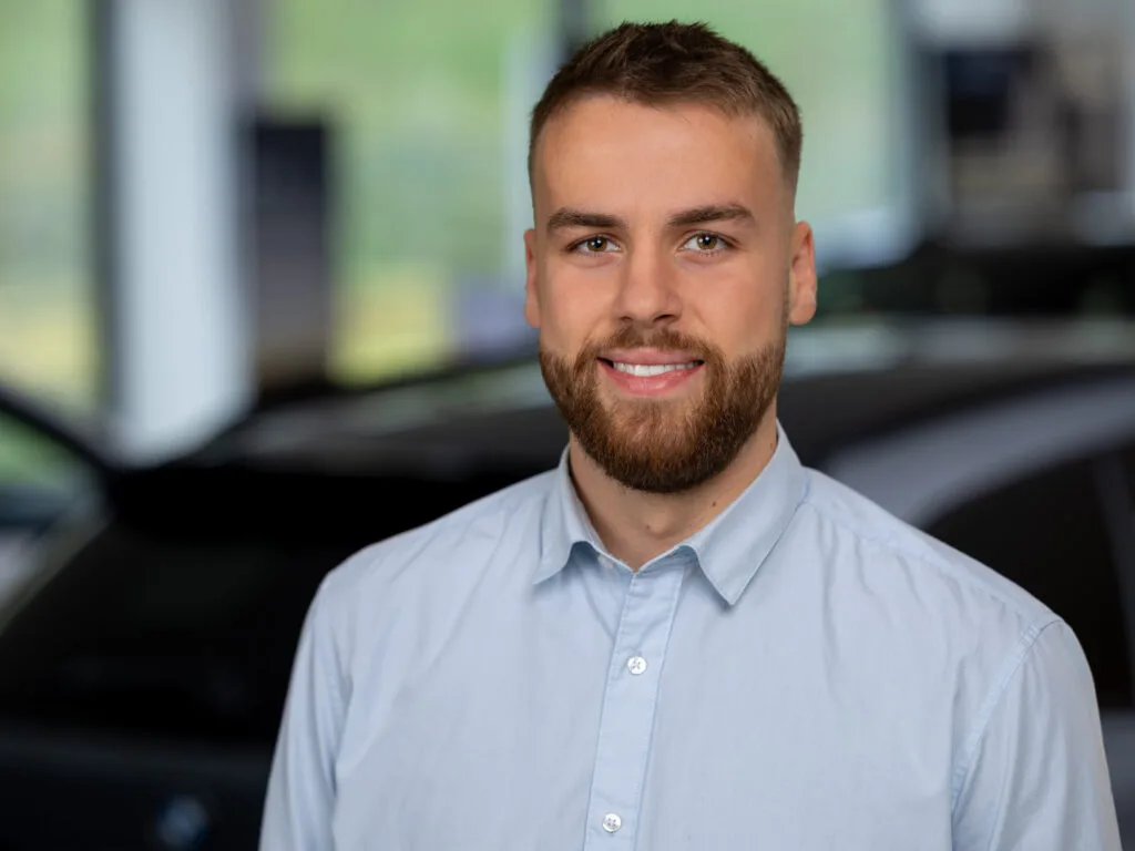 Kaltenbach Gruppe – Ein junger Mann mit kurzen braunen Haaren und einem Bart lächelt in die Kamera. Er trägt ein hellblaues Button-up-Hemd und steht in einem Haus in Kaltenbach-Remscheid. Im Hintergrund ist unscharf ein dunkles Auto zu sehen. – BMW, MINI, KIA, BMW Motorrad & Honda Motorrad Partner in NRW