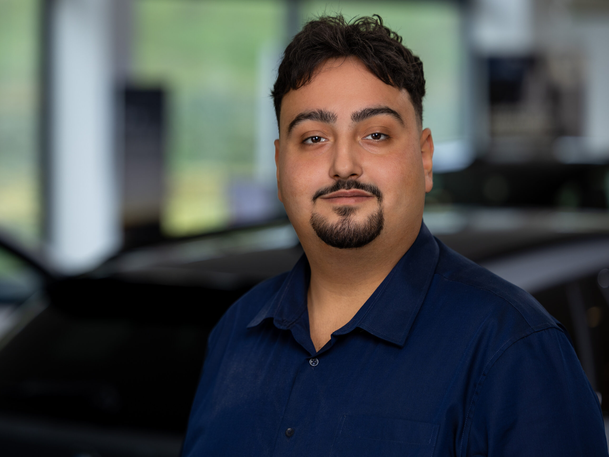 Kaltenbach Gruppe – Ein Mann mit kurzen dunklen Haaren und gestutztem Bart, der ein marineblaues Hemd trägt, steht in einem Haus vor einem unscharfen Hintergrund, in dem ein schwarzes Auto in Kaltenbach-Werdohl zu sehen ist. – BMW, MINI, KIA, BMW Motorrad & Honda Motorrad Partner in NRW