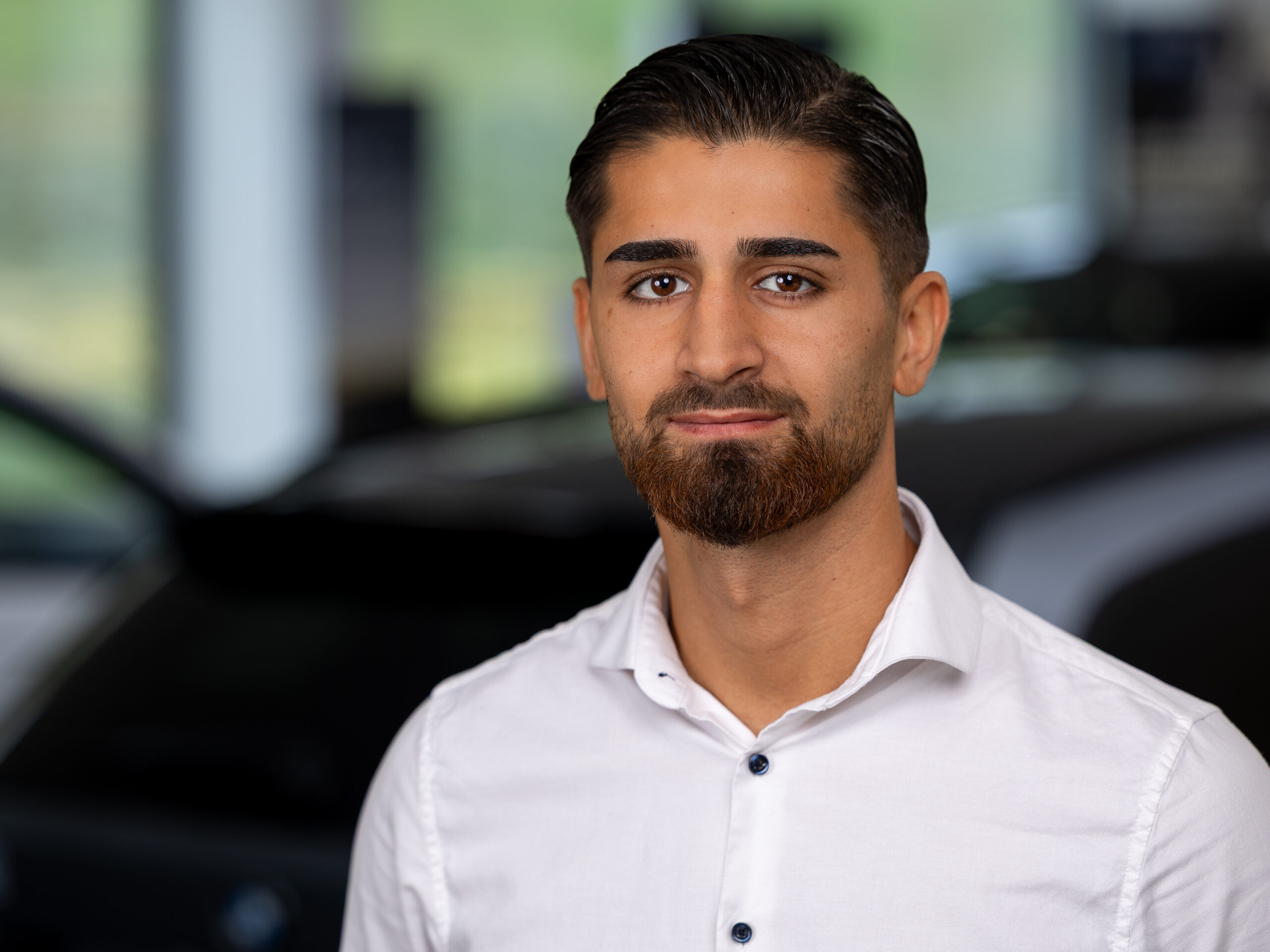 Kaltenbach Gruppe – Ein junger Mann mit kurzen dunklen Haaren und gestutztem Bart, der ein weißes Button-up-Hemd trägt, steht in einem Innenraum von Kaltenbach Olpe. Der unscharfe Hintergrund lässt auf ein Büro oder einen Ausstellungsraum schließen. – BMW, MINI, KIA, BMW Motorrad & Honda Motorrad Partner in NRW