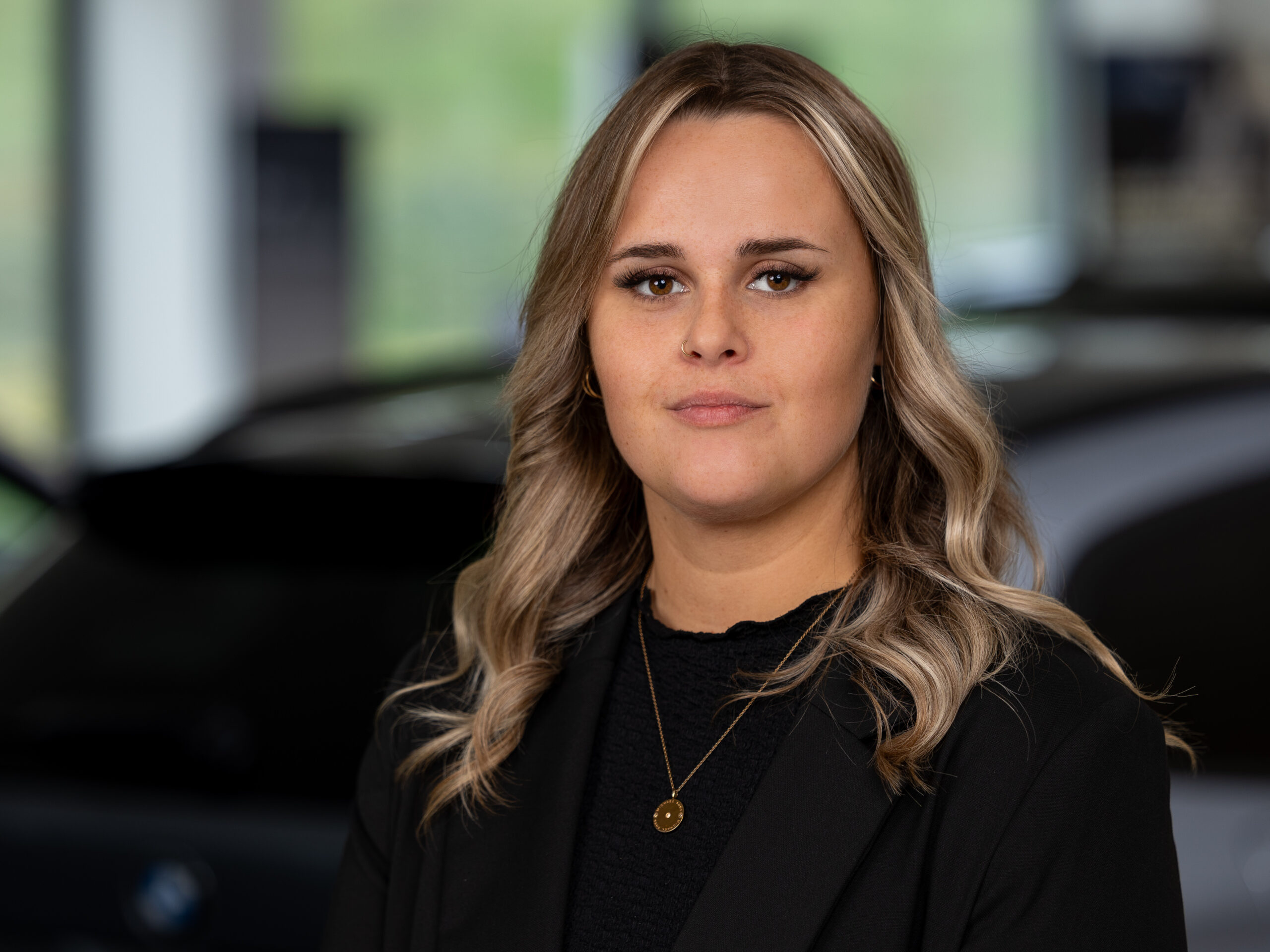 Kaltenbach Gruppe – Eine Frau mit langen, gewellten blonden Haaren, einem schwarzen Blazer und einer Halskette mit Anhänger steht in einem Innenraum in Kaltenbach Olpe, vor einem unscharfen Hintergrund mit einem Auto. – BMW, MINI, KIA, BMW Motorrad & Honda Motorrad Partner in NRW