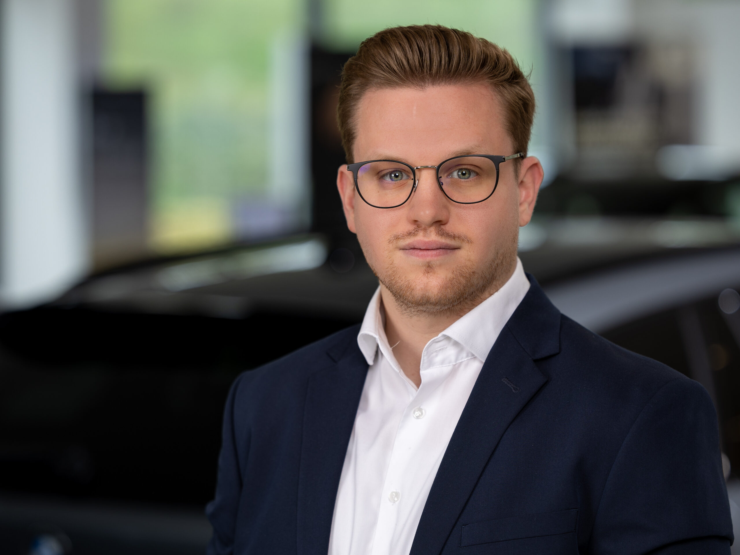 Kaltenbach Gruppe – Ein Mann mit kurzem, gestyltem Haar und Brille, der eine marineblaue Anzugsjacke und ein weißes Hemd trägt, steht in einem Innenraum vor einem unscharfen Hintergrund mit einem Auto in Kaltenbach Lennestadt. – BMW, MINI, KIA, BMW Motorrad & Honda Motorrad Partner in NRW