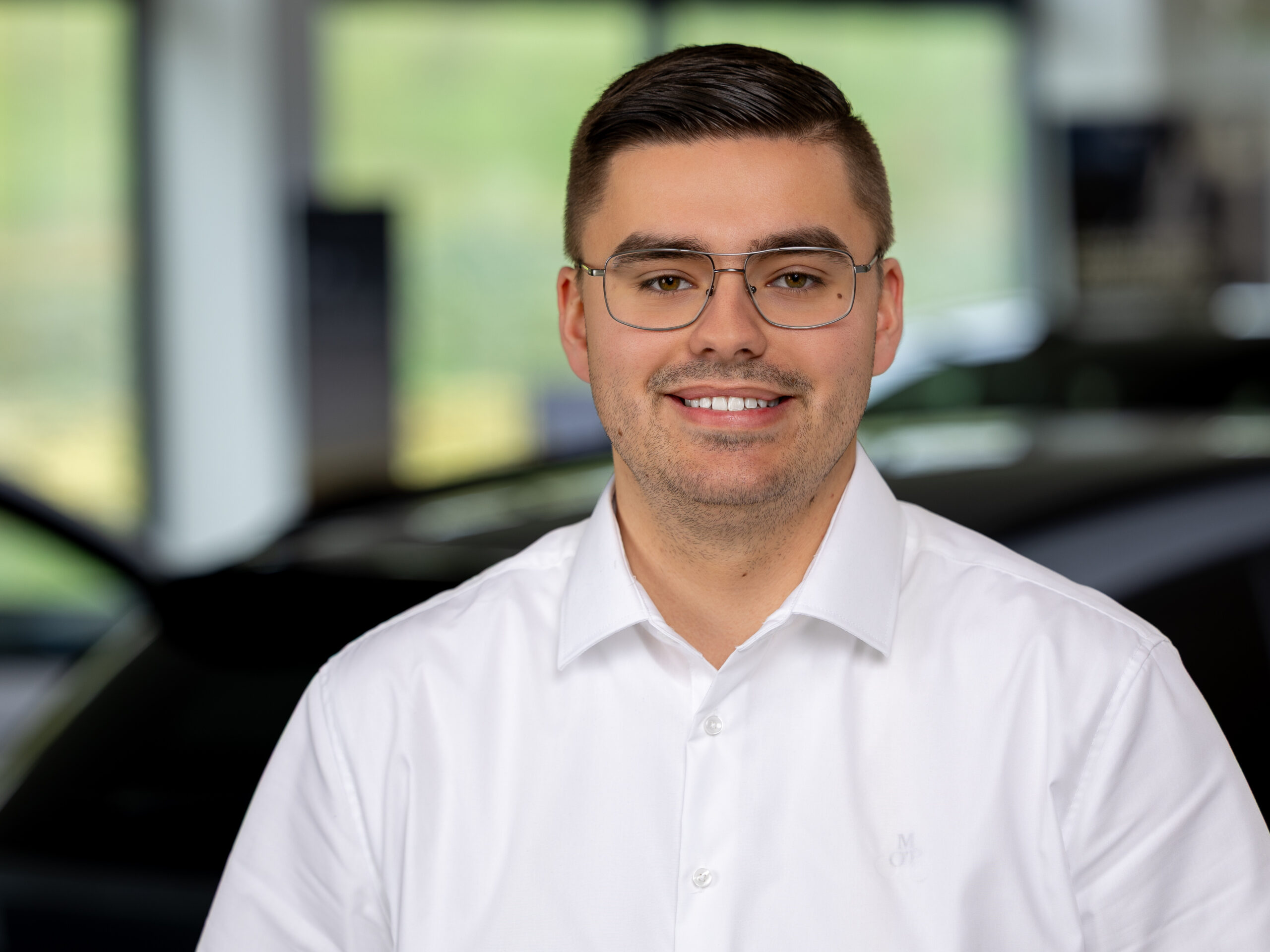 Kaltenbach Gruppe – Ein Mann mit kurzen dunklen Haaren und Brille, der ein weißes Hemd trägt, lächelt in die Kamera. Der unscharfe Hintergrund suggeriert einen Innenraum in Kaltenbach Overath, in dem Fenster und ein dunkles Auto zu sehen sind. – BMW, MINI, KIA, BMW Motorrad & Honda Motorrad Partner in NRW