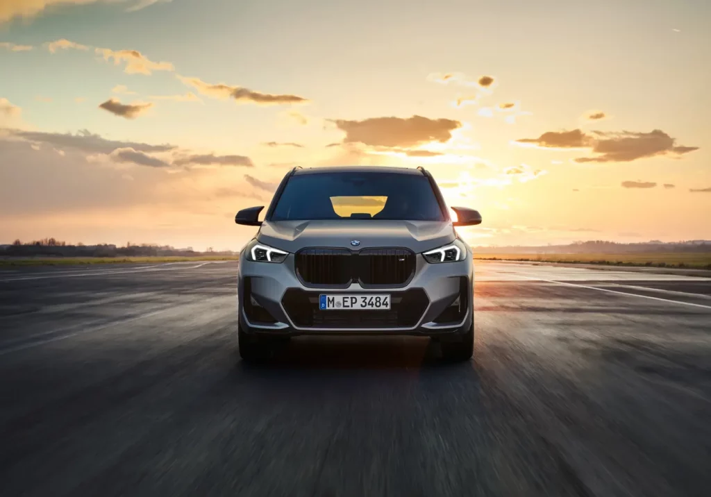 Kaltenbach Gruppe – Frontansicht eines silbernen BMW-Geländewagens, der bei Sonnenuntergang auf einer offenen Straße fährt, wobei die Sonne tief am Horizont steht und die Wolken am Himmel verstreut sind. – BMW, MINI, KIA, BMW Motorrad & Honda Motorrad Partner in NRW