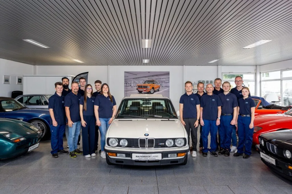 Kaltenbach Gruppe – Eine Gruppe von dreizehn Personen in passenden marineblauen Hemden steht im Ausstellungsraum von Kaltenbach um einen BMW Classic herum, während im Hintergrund weitere Autos und Autodekoration zu sehen sind. – BMW, MINI, KIA, BMW Motorrad & Honda Motorrad Partner in NRW