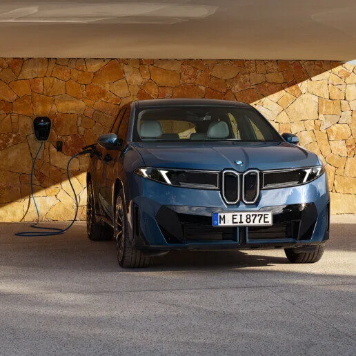 Kaltenbach Gruppe – Ein blaues BMW iX3-Elektroauto wird an einer Wandladestation vor einer Steinmauer in einem schattigen Bereich geparkt und aufgeladen. – BMW, MINI, KIA, BMW Motorrad & Honda Motorrad Partner in NRW
