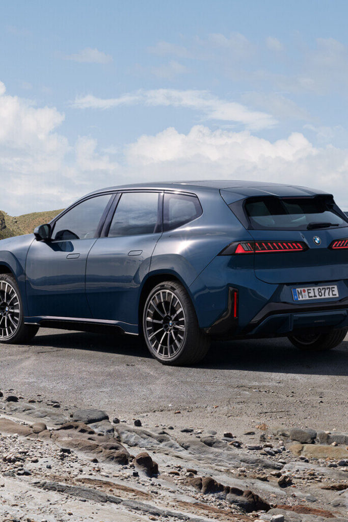 Kaltenbach Gruppe – Ein dunkelblauer BMW iX3 SUV ist auf einer gepflasterten Fläche in der Nähe eines felsigen Ufers geparkt, mit Hügeln und einem teilweise bewölkten Himmel im Hintergrund. Das Fahrzeug wird von der Rückseite aus betrachtet. – BMW, MINI, KIA, BMW Motorrad & Honda Motorrad Partner in NRW