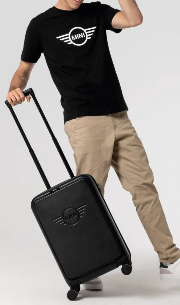 Kaltenbach Gruppe – Eine Person, die ein schwarzes MINI-T-Shirt und eine beigefarbene Hose trägt, steht mit dem Griff eines schwarzen Rollkoffers mit dem MINI-Logo in der Hand vor einem schlichten hellen Hintergrund. – BMW, MINI, KIA, BMW Motorrad & Honda Motorrad Partner in NRW