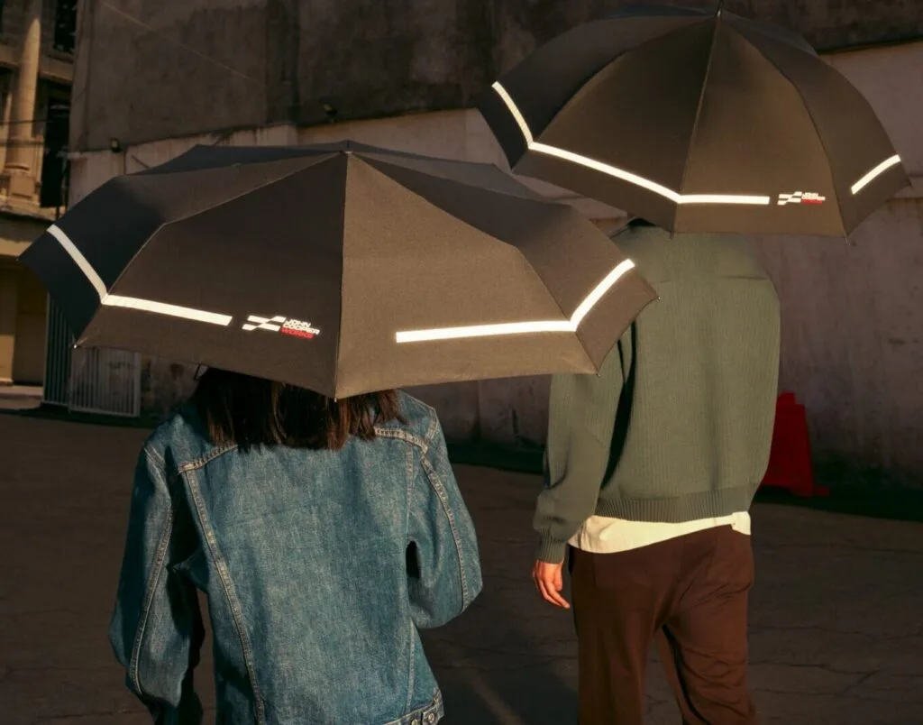 Kaltenbach Gruppe – Zwei Personen, eine in Jeansjacke und heller Hose und die andere in grüner Jacke und brauner Hose, gehen im Freien mit schwarzen Regenschirmen mit reflektierenden Streifen auf einer gepflasterten Fläche in der Nähe einer Betonwand. – BMW, MINI, KIA, BMW Motorrad & Honda Motorrad Partner in NRW