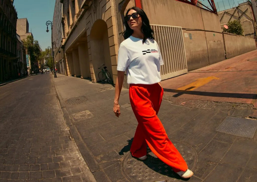 Kaltenbach Gruppe – Eine Frau mit Sonnenbrille, weißem T-Shirt und leuchtend roter Hose geht selbstbewusst eine sonnenbeschienene Straße in der Stadt entlang, mit alten Gebäuden und einem geparkten Fahrrad im Hintergrund. – BMW, MINI, KIA, BMW Motorrad & Honda Motorrad Partner in NRW
