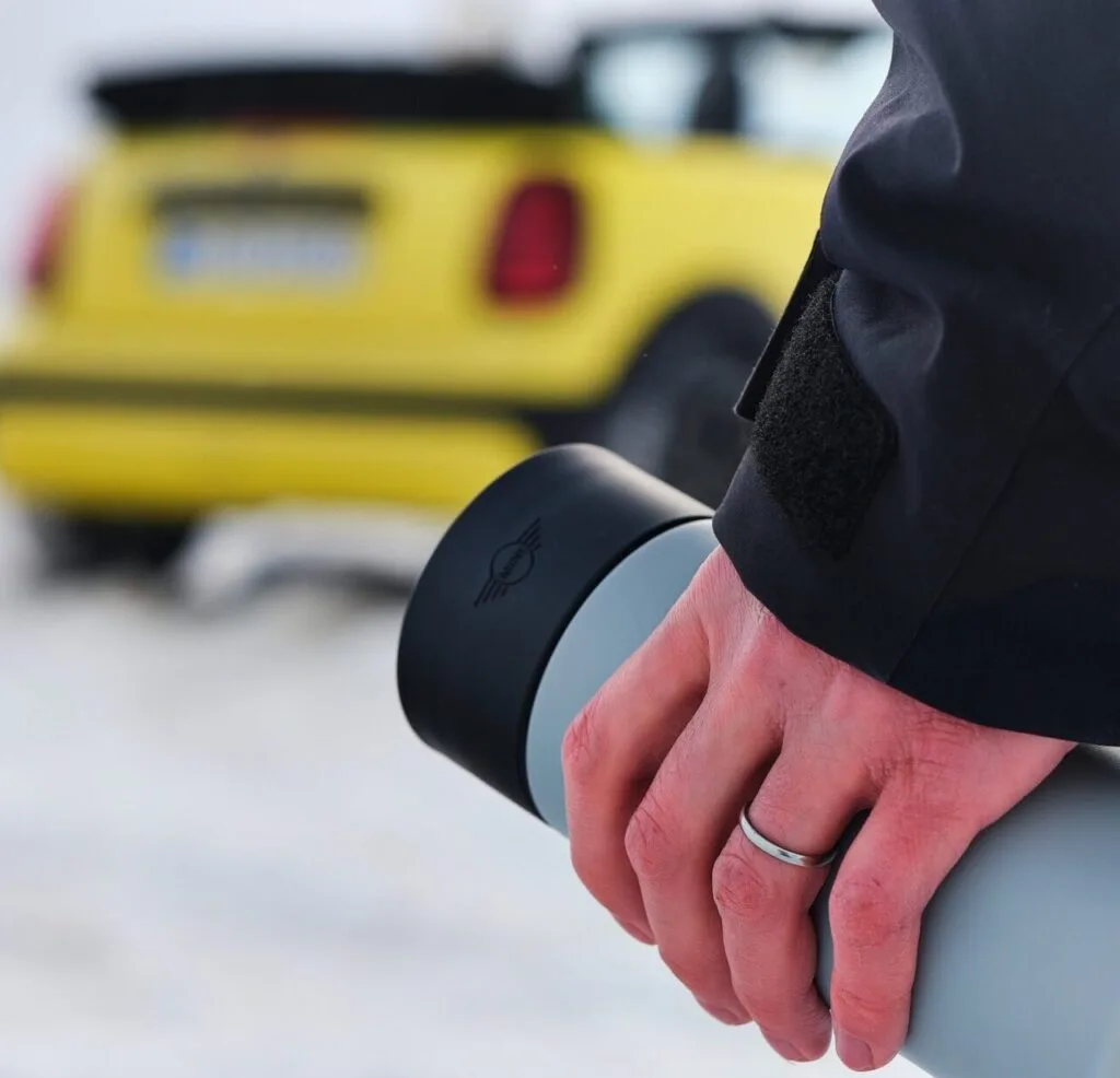 Kaltenbach Gruppe – Nahaufnahme einer Person, die eine graue Thermosflasche hält, mit einem gelben Auto, das im verschneiten Hintergrund geparkt ist. Die Person trägt eine dunkle Jacke, und ihr Ehering ist sichtbar. – BMW, MINI, KIA, BMW Motorrad & Honda Motorrad Partner in NRW