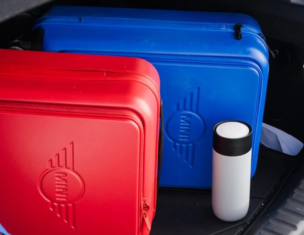 Kaltenbach Gruppe – Zwei Hartschalenkoffer, ein roter und ein blauer, liegen im Kofferraum eines Autos neben einem weißen Reisebecher. Auch ein Teil eines Nummernschilds und eine blaue Tasche sind zu sehen. – BMW, MINI, KIA, BMW Motorrad & Honda Motorrad Partner in NRW