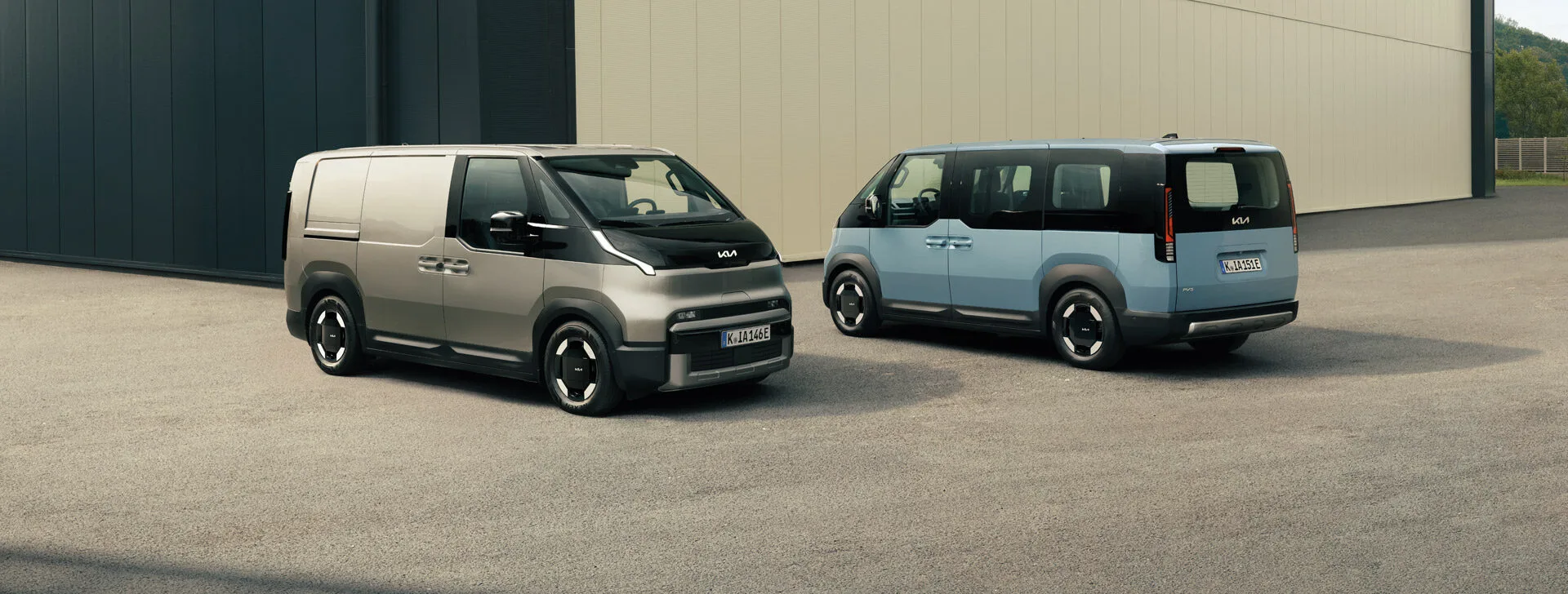 Der Kia PV5 Cargo in schräger Vorderansicht und der Kia PV5 Passenger in schräger Hinteransicht zusammen vor einem gewerblichen Gebäude